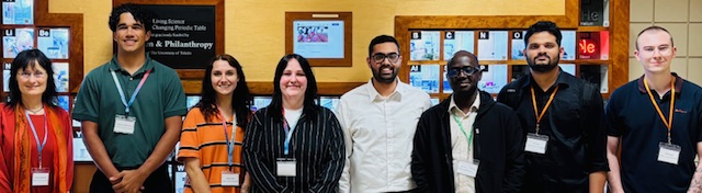 Eight members of the Pohl lab standing in front of a periodic table at the University of Toledo